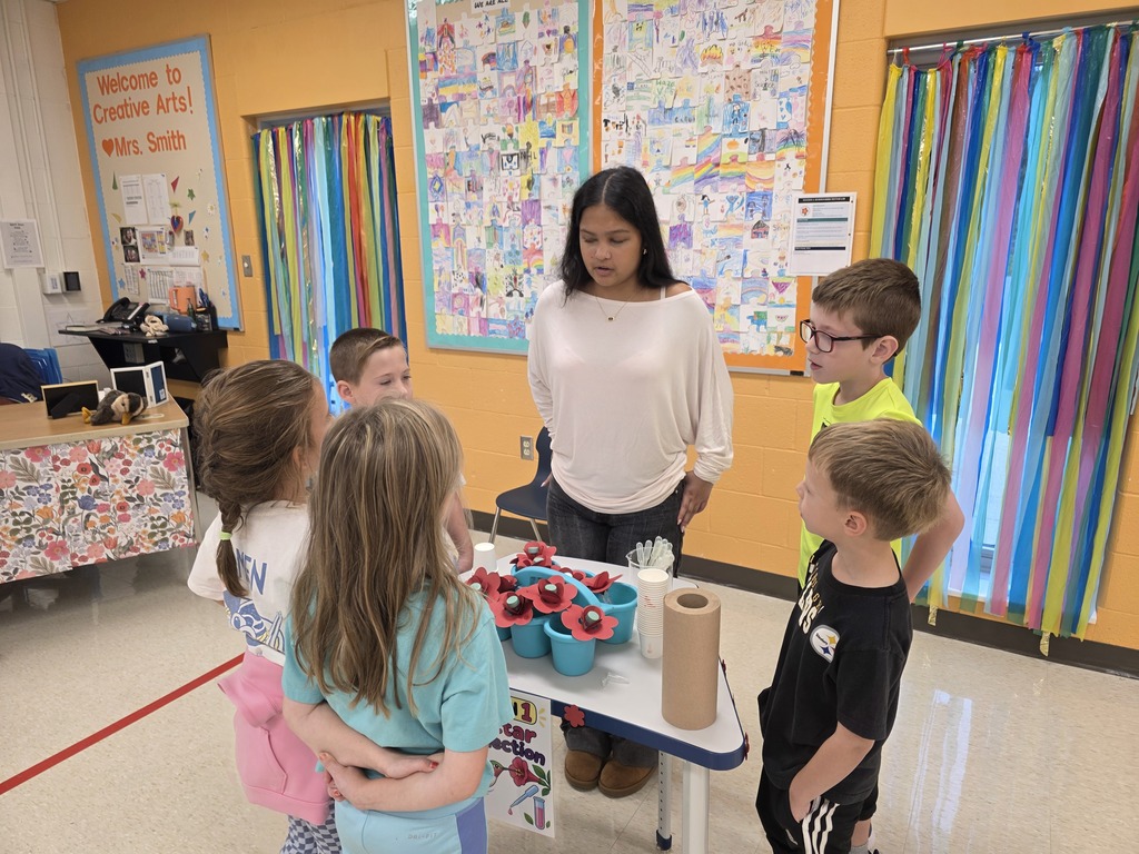 A woman in a white shirt stands with children around a table with blue bowls and a roll of paper.