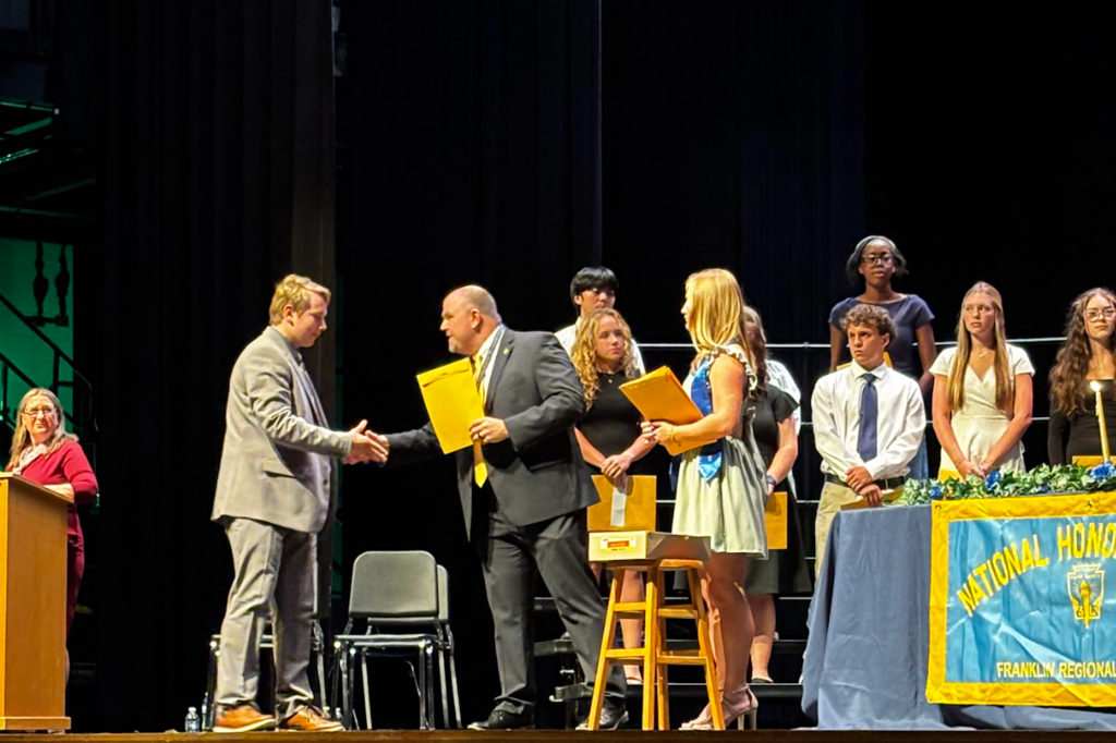 A stage with people in suits and dresses, possibly during an award ceremony. One person is holding a folder.
