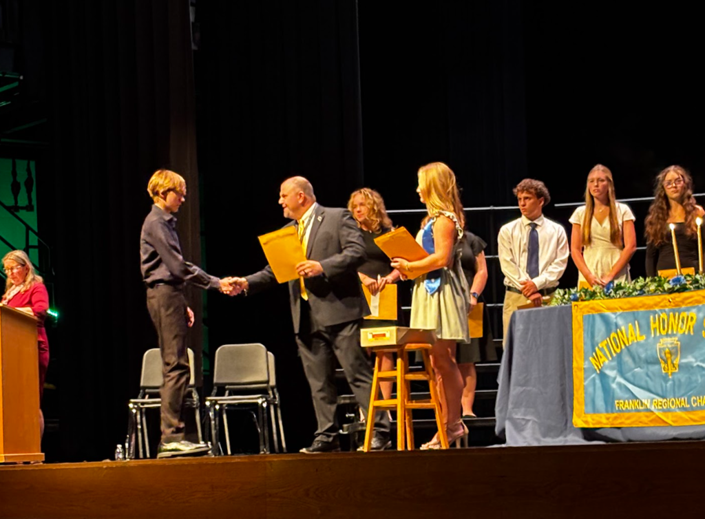 Group of people on stage with two individuals shaking hands. One holds a certificate. Banner behind them.