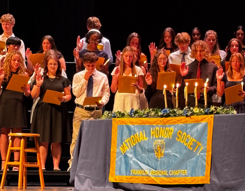 A group of students on stage at an event, holding books, some raising their hands. A table displays a banner for the National Honor Society.