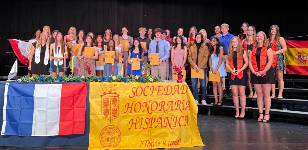 Group of students on stage with podium banner reading "Sociedad Honoraria Hispanica". French and Spanish flags on table.