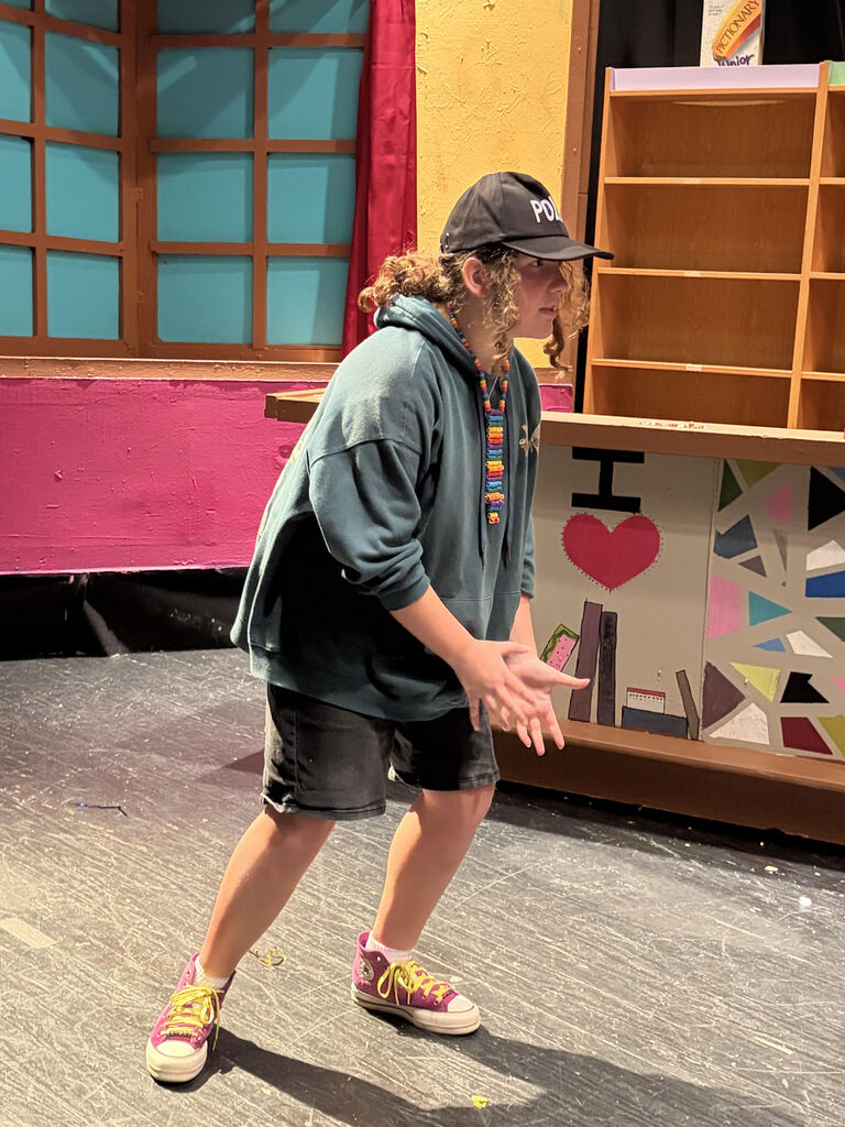 Person wearing a baseball cap, hoodie, and sneakers on a stage, gesturing with hands.