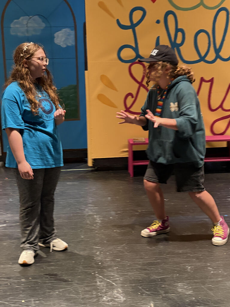 Two individuals on stage, one in blue and one in green, face each other. Behind them is a vibrant backdrop with the words "Likely" and a pink bench.