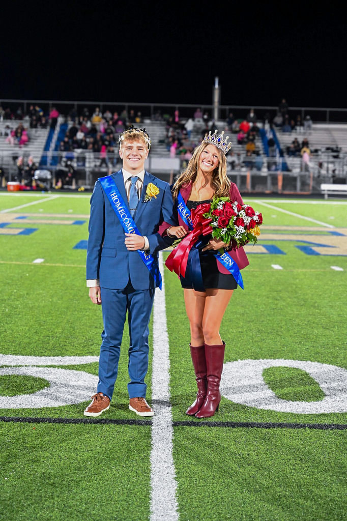 A man and a woman stand on a football field. They are both wearing crowns. The woman holds flowers.