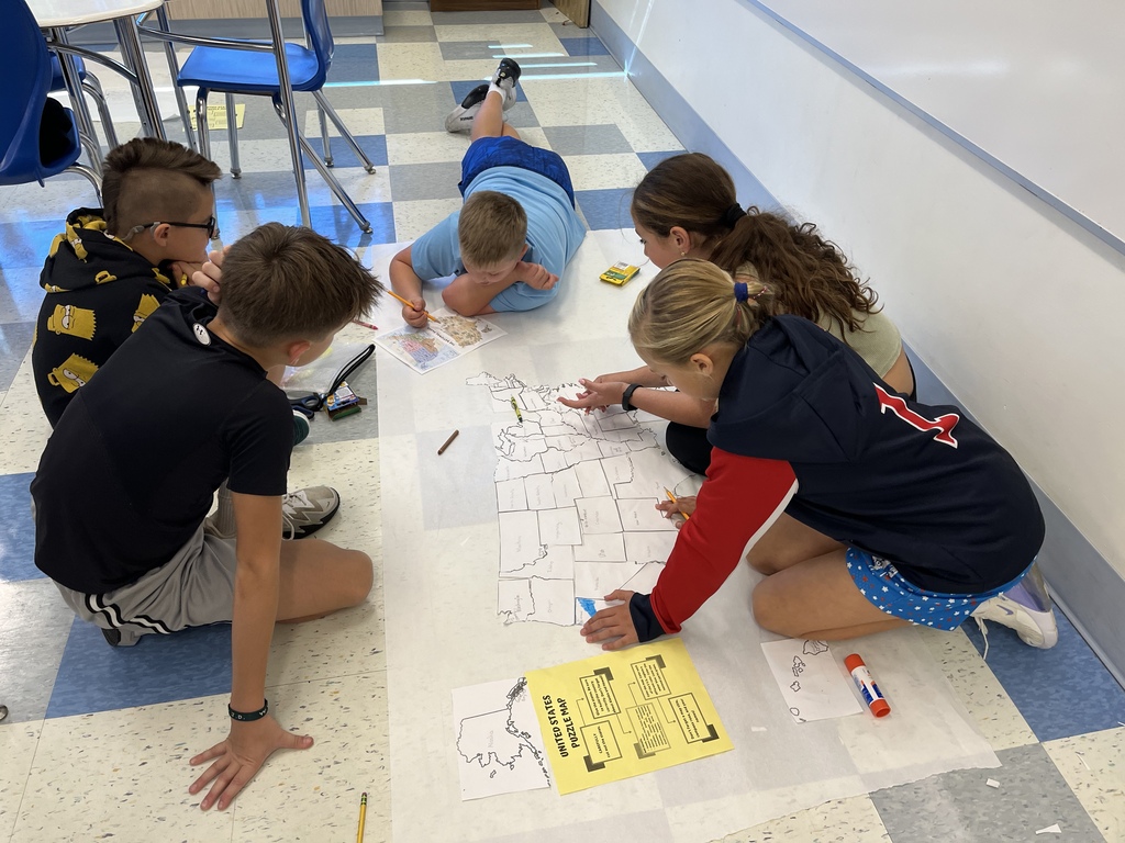 Five children on floor in classroom. Three are kneeling, one lying down, and one standing, working on a large map on the floor.