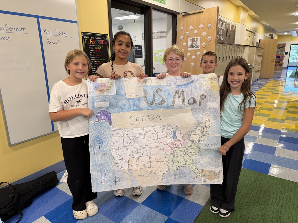 Five children in a classroom stand behind a large poster of a U.S. map. Two children hold the map.