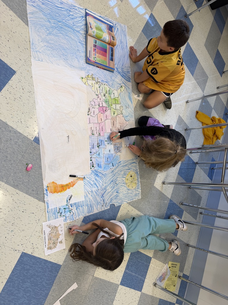 Three children work on a large map, one lying down, another kneeling, and the third seated, with crayons and a book nearby.