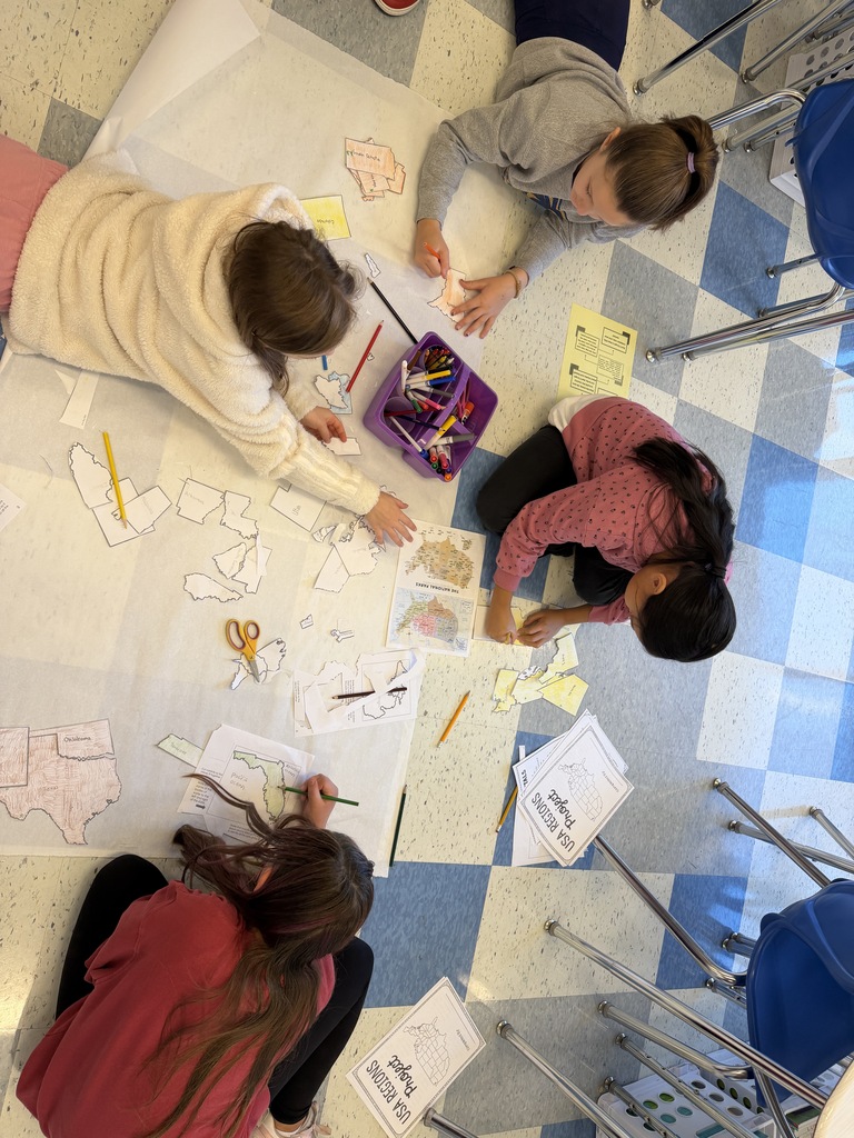 Four children work on a large project on a floor, surrounded by papers, scissors, and pencils.