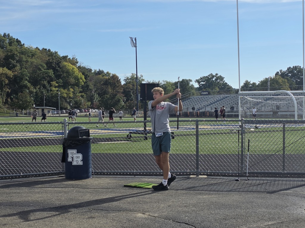 A man on a field holding a pole, possibly practicing a sport. A trash can is nearby.