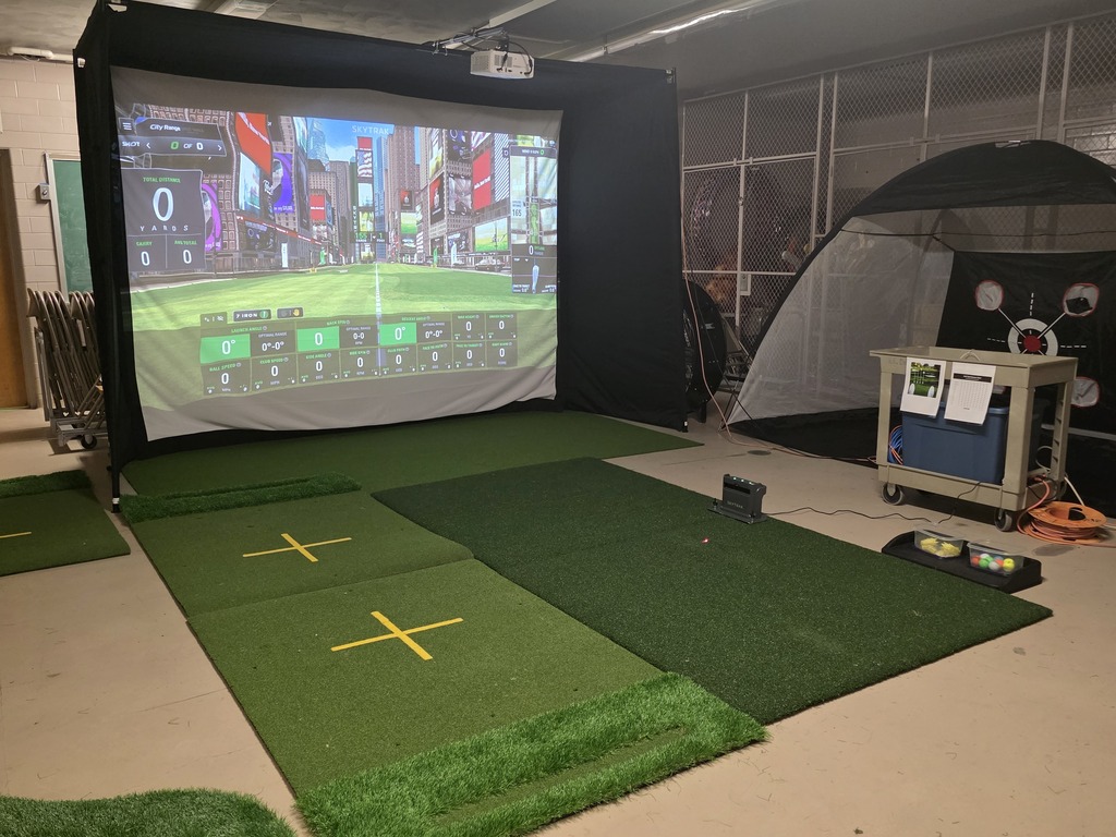 Indoor golf simulator setup with projector screen, green mats, and a tent, set against a white wall.