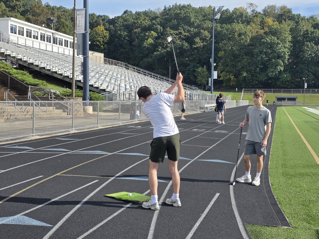 Two men play golf on a track, one swinging a club, another holding a putter, with empty bleachers and trees in the background.