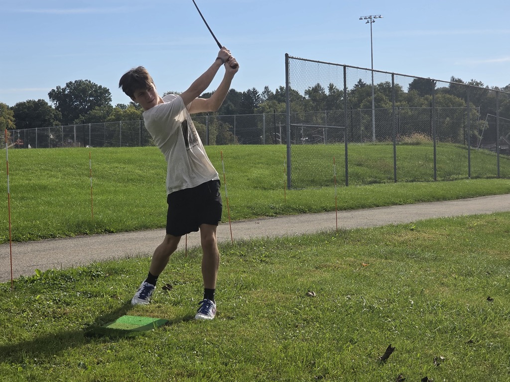 A person swings a golf club in a park, with a grassy field, a fence, and trees in the background.