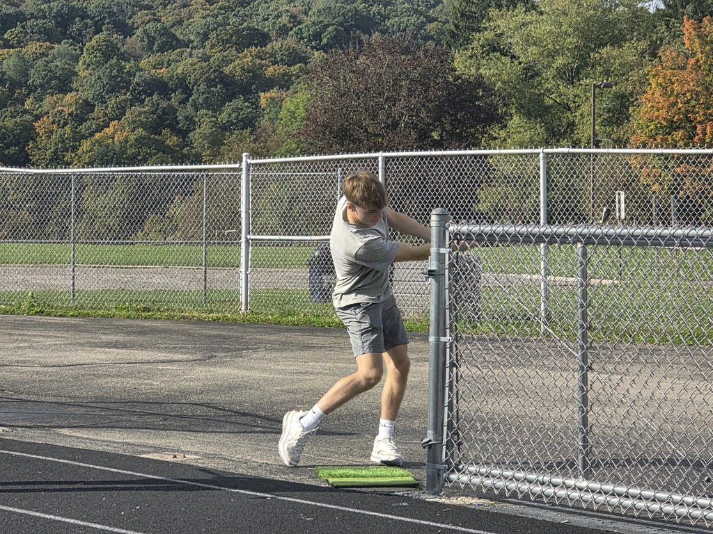 An individual swings a hammer at an outdoor sports venue. Behind the fence, lush green trees surround a grassy field.