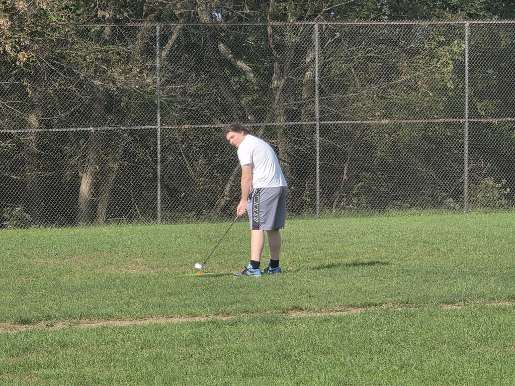 A person in a white shirt and gray shorts plays golf on a grassy field, with a chain-link fence and trees in the background.