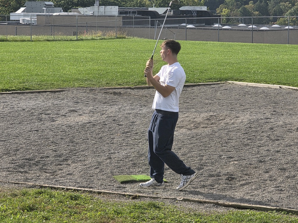 A man practices golf swings on a sandy surface with a green field and fence in the background.