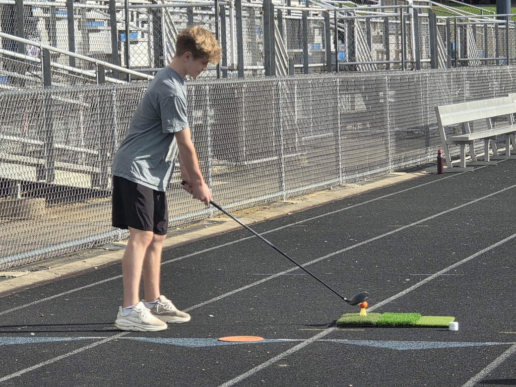 A young person practices golf on a track with a metal fence, a bench, and an orange circle.
