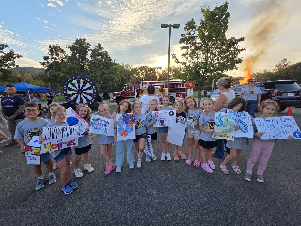 Kids stand in a line with signs in a parking lot, a fire truck is visible in the background.