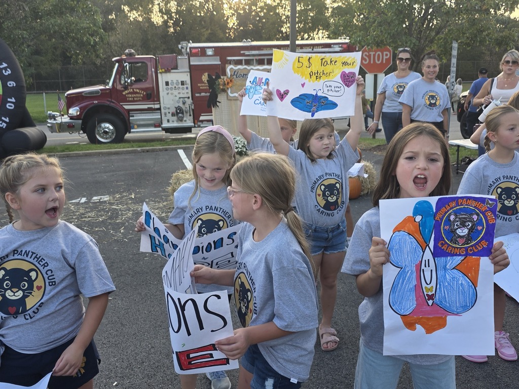 Children with gray shirts holding signs, including butterflies, walk on a street. Firetruck and people in background.