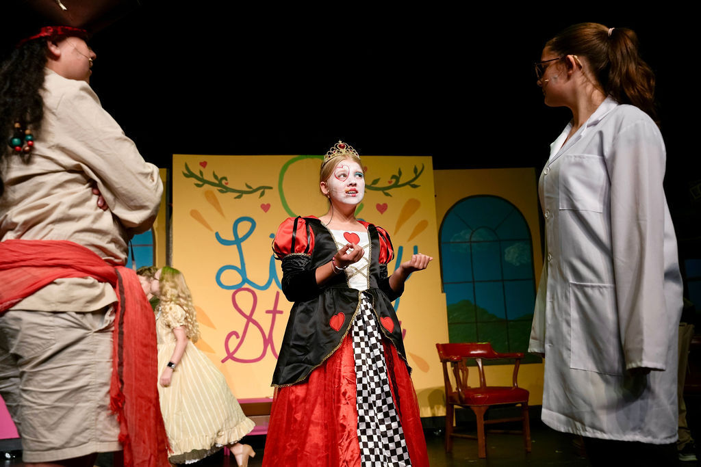 Three performers on stage; one in a red and white checkered dress, another in a white coat, and the third in a tan outfit.