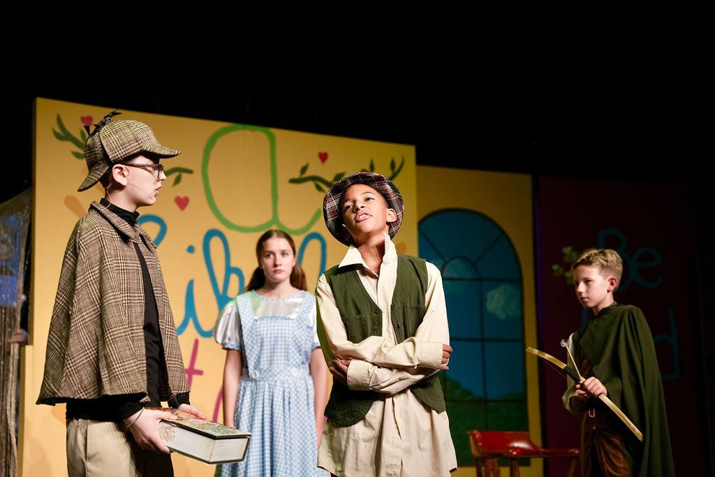 Four individuals in costumes on a stage, one holding a book, another with a stick, and a backdrop featuring multicolored sections.