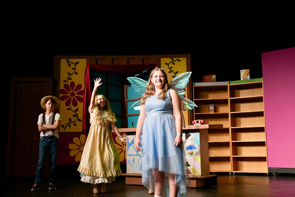 Three children in costumes perform on a stage. One child wears fairy wings, another a yellow dress.