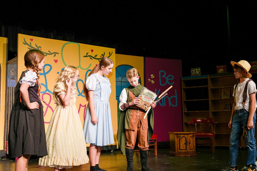 Five children on stage, four girls and one boy, wearing costumes, with colorful backdrops and a wooden box.