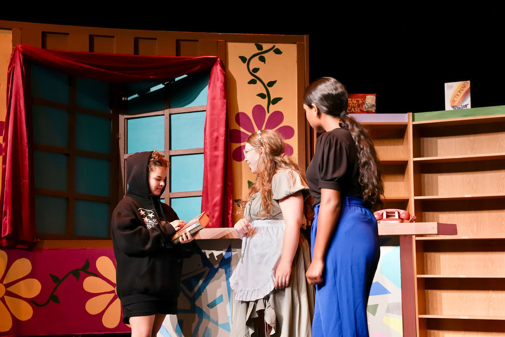 Three individuals stand before a colorful backdrop with shelves and flowers. One holds an object, while the other two look at it.
