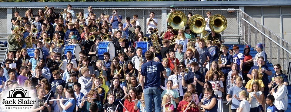 A large group of people in a stadium, some playing instruments, including brass and percussion, with a blue banner visible.