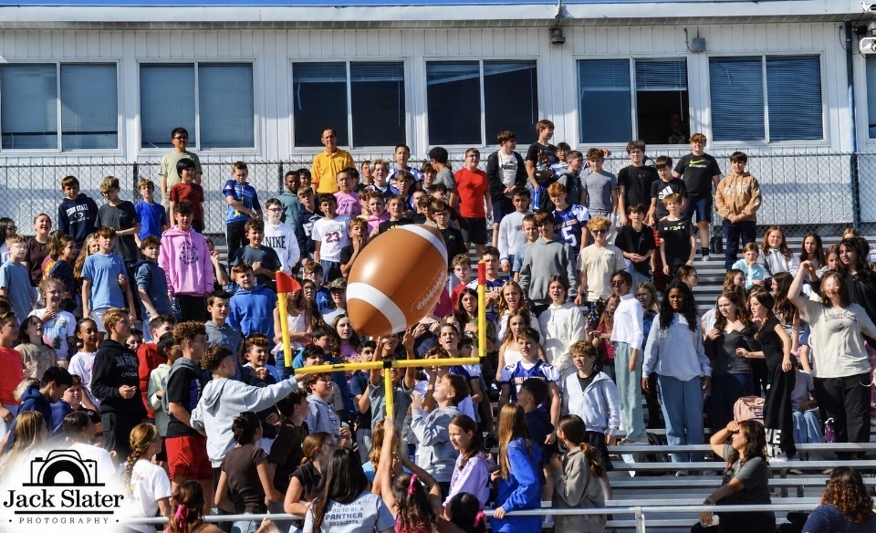 A crowd of people in stands at a football event, cheering. A football on a yellow goalpost is lifted.