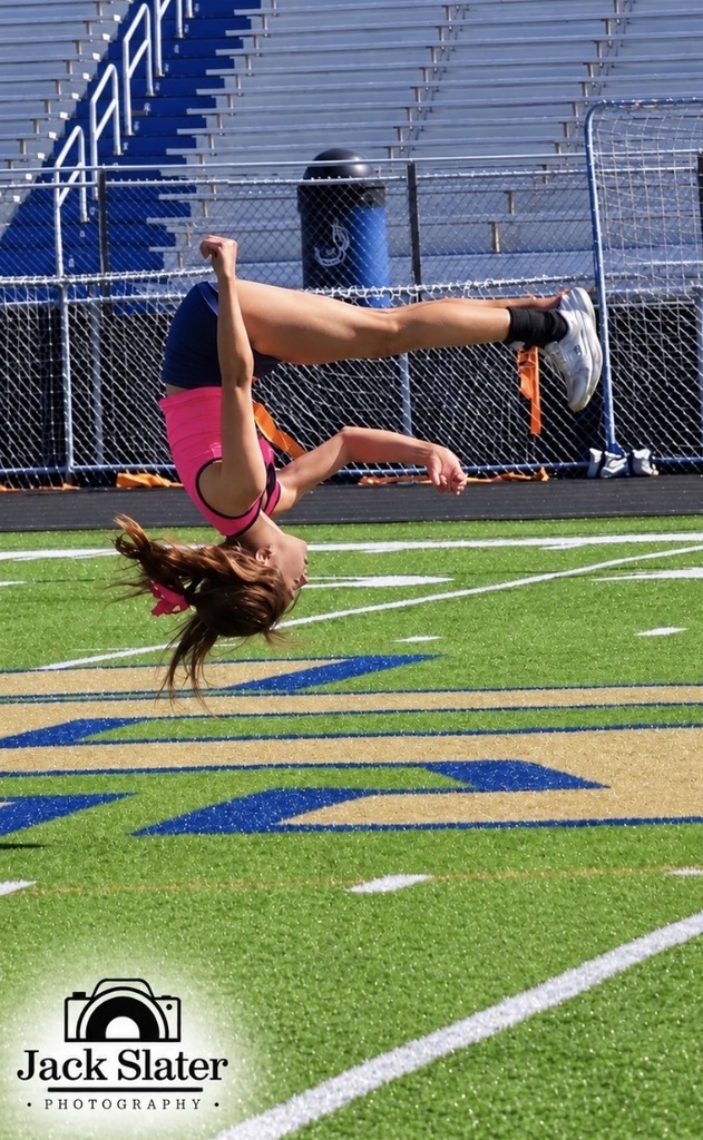 A female gymnast performs a handstand on a field, with blue bleachers and a blue trash bin in the background.