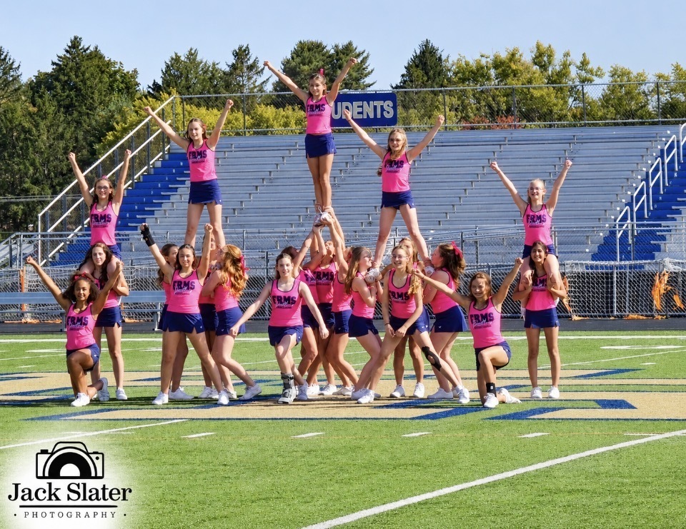 Cheerleaders in pink and blue uniforms form a pyramid on a football field with bleachers and trees in the background.