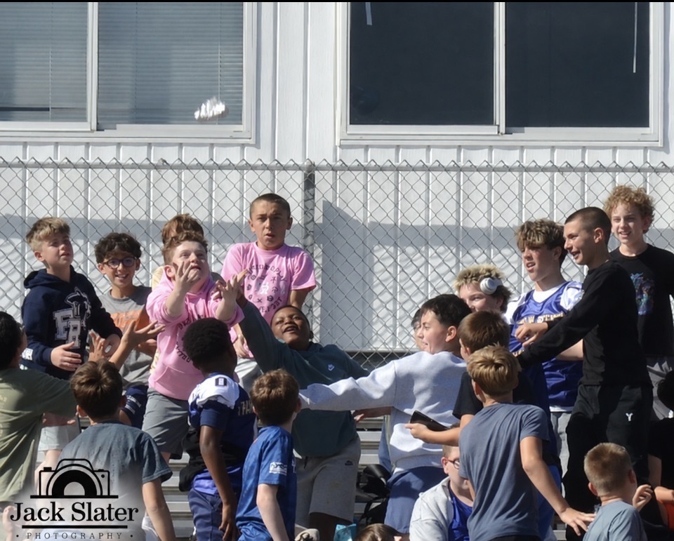 Group of kids on bleachers, some wearing sports jerseys, near a fence with windows.