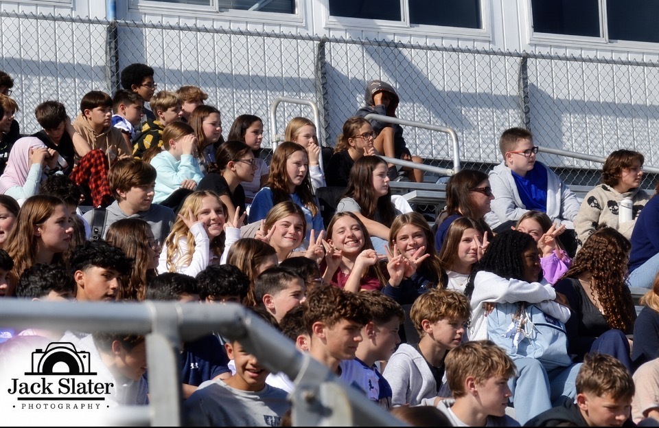 A large group of people, possibly students, sit on bleachers in an outdoor stadium. Some make gestures.
