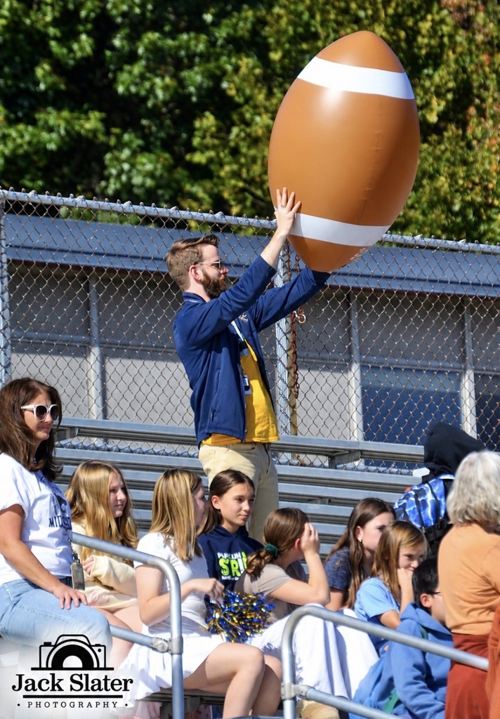 A man holds up an inflated football in a stadium, surrounded by spectators. Green trees are visible behind the fence.