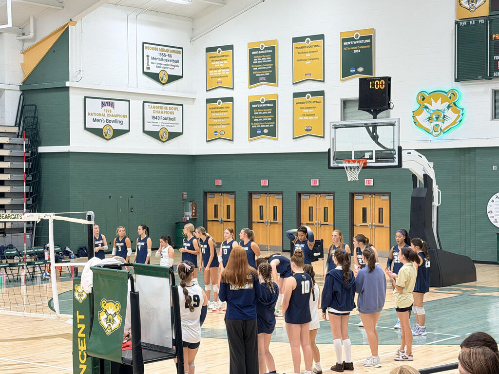 An indoor gym with green walls and wooden doors. A group of women in sports uniforms are gathered around a coach.
