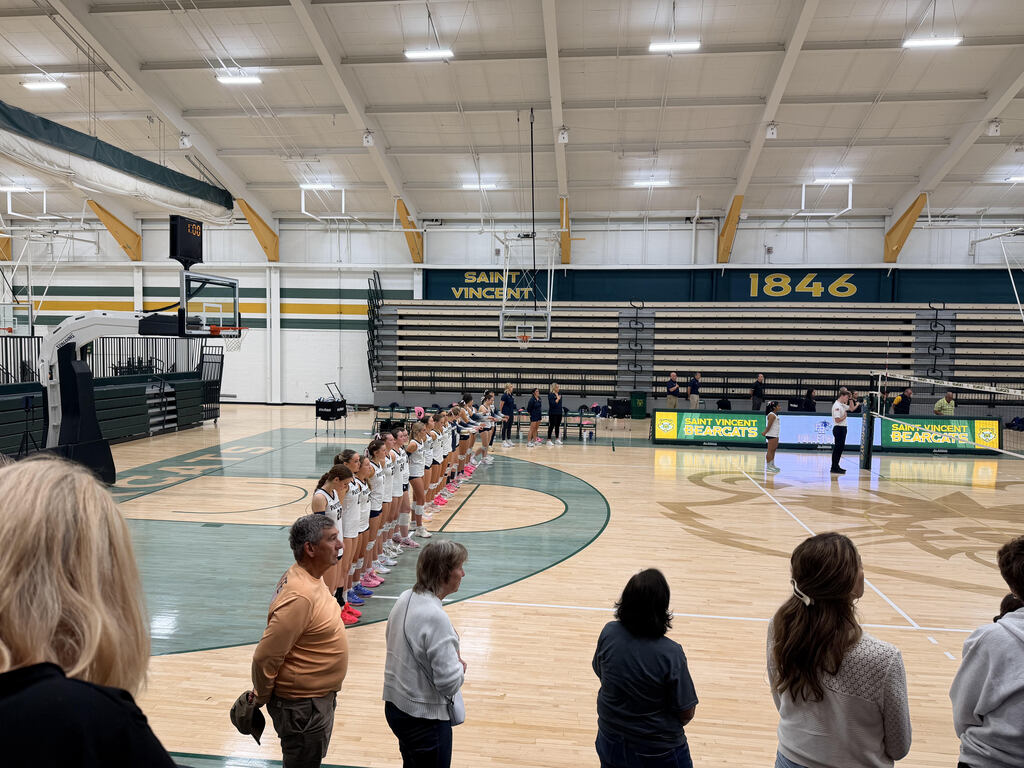 An indoor basketball court with players lined up. Spectators stand on the sidelines. Bright lights and year 1846 on the wall.