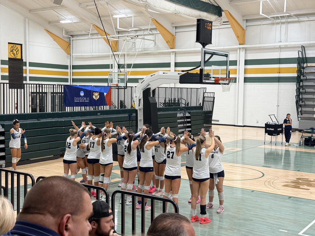 Volleyball team huddling on a court, facing the audience. Spectators seated on one side.