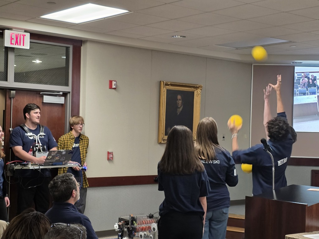 A group of people in blue shirts, one holding a yellow ball, standing before a podium in a room.