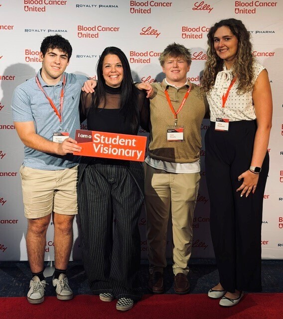 Four individuals stand on a red carpet. They wear lanyards with "Blood Cancer United" and "Student Visionary" signs.