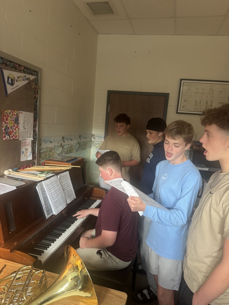 Five young males standing around a piano, one playing while the others observe and one holding music.
