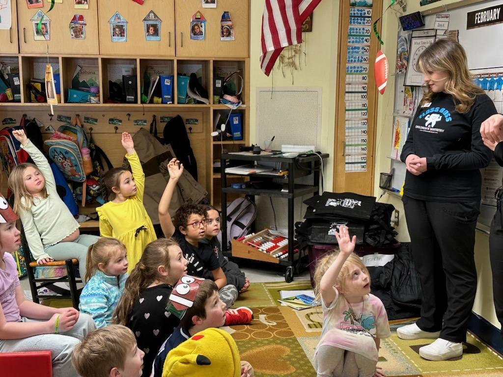 In a classroom, a group of children sit on the floor, listening to a woman. She stands and gestures while they raise their hands.