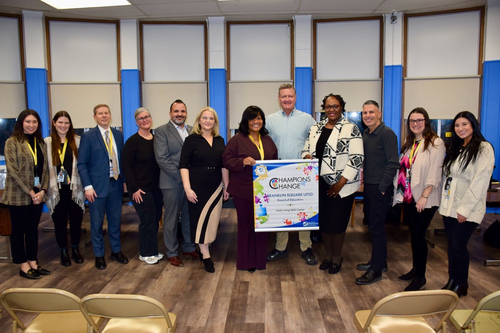 A diverse group of eleven people stands in a room, smiling and holding a colorful "Champions of Change" banner. The atmosphere is celebratory and formal.