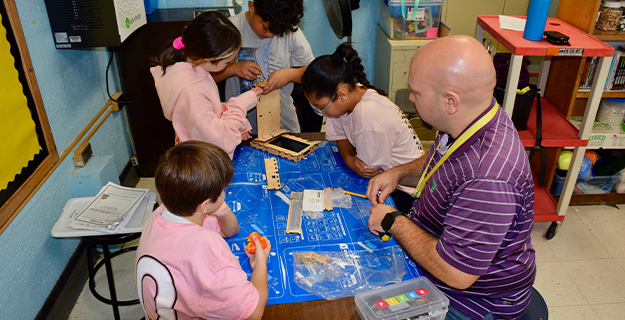 Teacher sitting with students at blue desk.