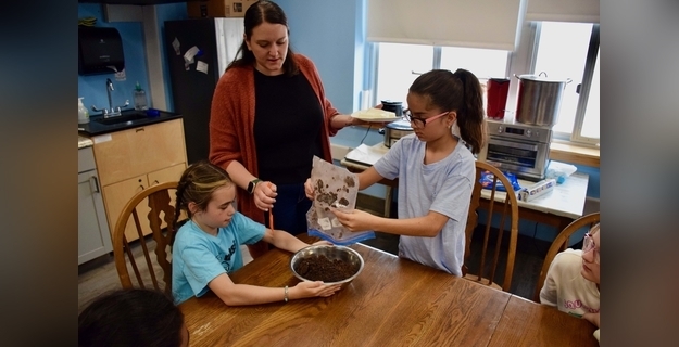Children learning to mix soil for a gardening project with an instructor in a bright, inviting kitchen.
