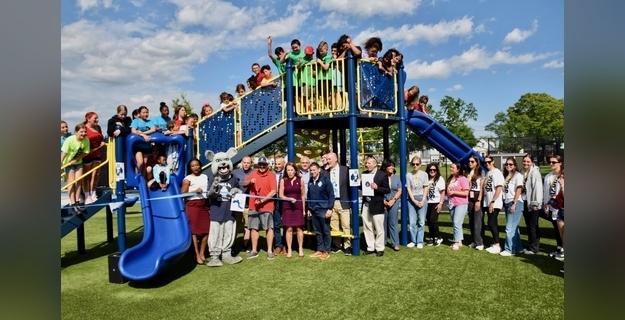 Children and adults celebrate the grand opening of a new playground, featuring a blue slide and climbing structure, with a mascot present among the crowds.