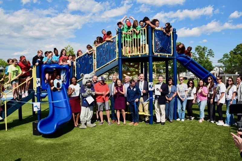 Community celebration at a newly opened playground with students and adults gathered around a colorful play structure under a bright blue sky.