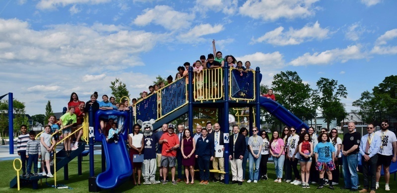 Community celebration at a newly opened playground with students and adults gathered around a colorful play structure under a bright blue sky.