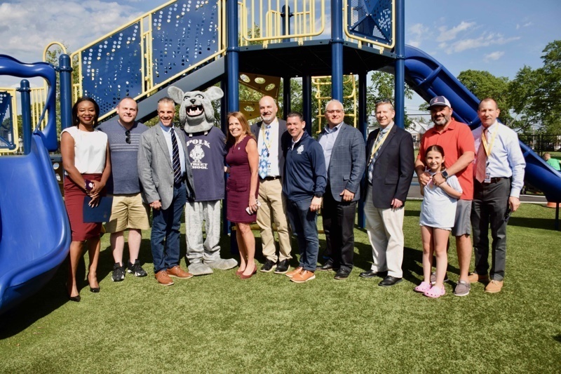 Community celebration at a newly opened playground with students and adults gathered around a colorful play structure under a bright blue sky.