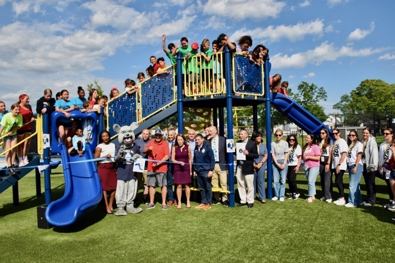 Community celebration at a newly opened playground with students and adults gathered around a colorful play structure under a bright blue sky.