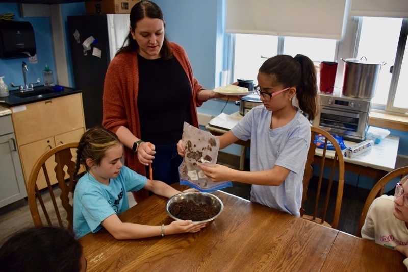 Children learning to mix soil for a gardening project with an instructor in a bright, inviting kitchen.